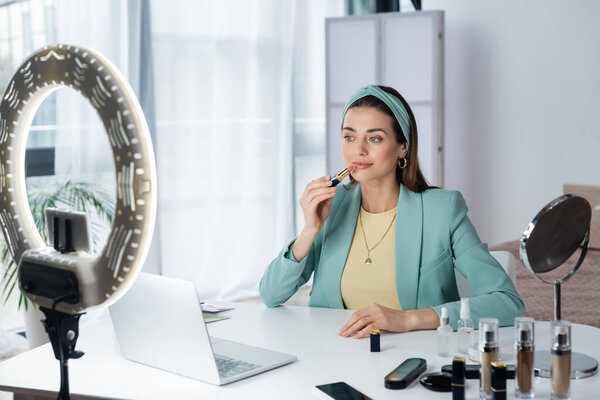 красивая женщина, применяющая помаду рядом с ноутбуком, смартфон с пустым экраном и круговой лампой