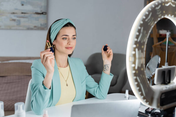 Блогер красоты, держащий помаду возле размытого ноутбука и держателя телефона с лампой