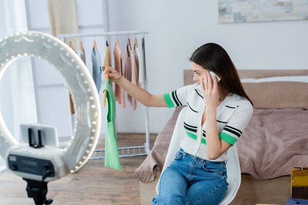 young woman holding hanger with vest while talking on smartphone near phone holder with ring lamp