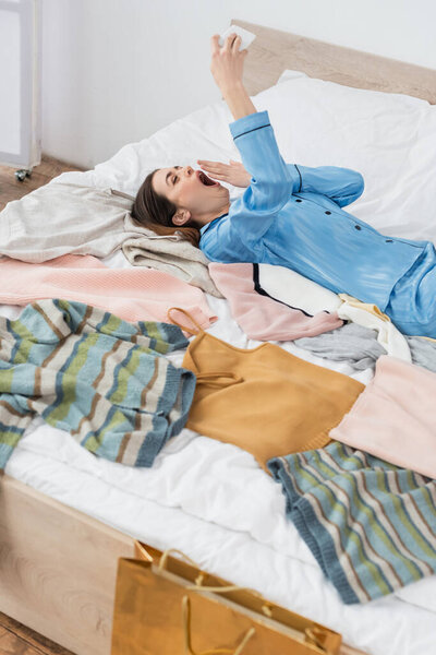 усталая женщина зевает во время видеозвонка рядом с большим количеством одежды на кровати