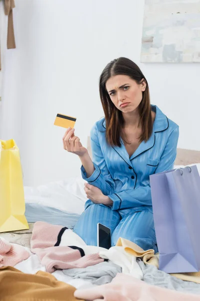 upset woman with credit card looking at camera near shopping bags and plenty of clothes on bed