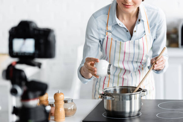 cropped view of culinary blogger adding salt into saucepan near blurred digital camera