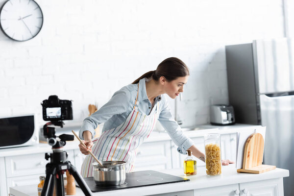 young woman looking at cookbook during food preparation in front of blurred digital camera