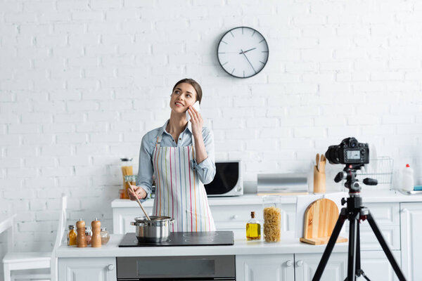 smiling culinary blogger talking on smartphone near saucepan and digital camera on tripod