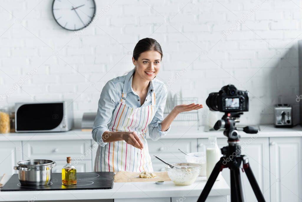 cheerful culinary blogger pointing with hand near dough and blurred digital camera