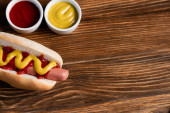 vrchní pohled na hořčici a kečup v miskách v blízkosti chutného hot dogu na dřevěném povrchu