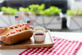 Hotdogs mit kleinen Fahnen, Schalen mit Senf und Ketchup in der Nähe karierter Servietten auf dem Tisch im Freien