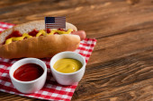 Schüsseln mit Soßen neben leckerem Hot Dog mit kleiner amerikanischer Flagge und karierter Serviette auf Holztisch