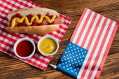 Draufsicht auf Hot Dog, Ketchup und Senf in der Nähe einer kleinen US-Flagge auf einem Holztisch