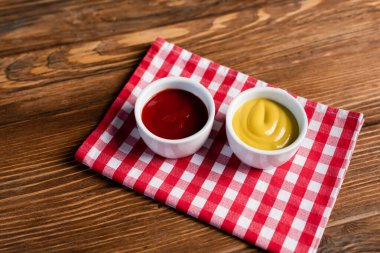 sauce bowls with ketchup and mustard on checkered napkin and wooden table clipart