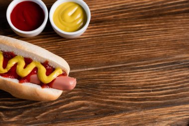 top view of mustard and ketchup in bowls near tasty hot dog on wooden surface clipart