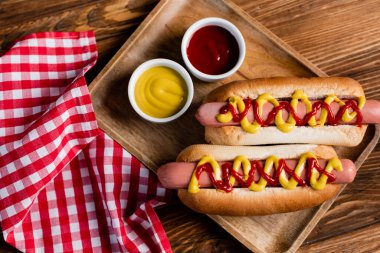 top view of hot dogs, ketchup and mustard near plaid napkin on wooden table clipart