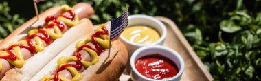 Küçük Amerikan bayraklı sosisliler, yeşil çimlerde ketçaplı ve hardallı kaselerin yanında pankart.
