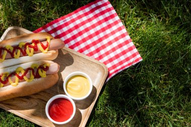 top view of hot dogs near sauces and plaid table napkin on green lawn clipart