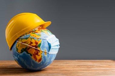medical mask and hardhat on globe on wooden surface isolated on grey, labor day concept clipart