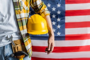Ekose gömlekli ve alet kemerli işçinin bulanık Amerikan bayrağının yanında şapka tutuşu, İşçi Bayramı konsepti.