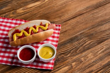bowls with ketchup and mustard near tasty hot dog on plaid table napkin and wooden surface clipart