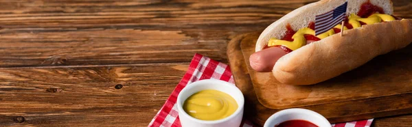 tasty hot dog with small usa flag near bowls with mustard and ketchup on wooden table, banner
