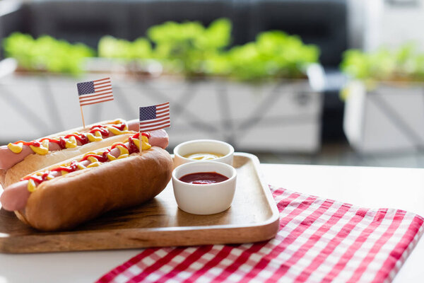 hot dogs with small us flags, bowls with mustard and ketchup near plaid napkin on table outdoors