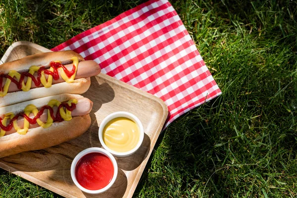 在绿色草坪上靠近酱料和铺好的餐巾纸的热狗的顶部视图 — 图库照片
