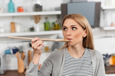 woman holding wooden spoon and trying food while cooking in kitchen  clipart