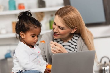 happy mother feeding adopted african american daughter with jello near laptop clipart