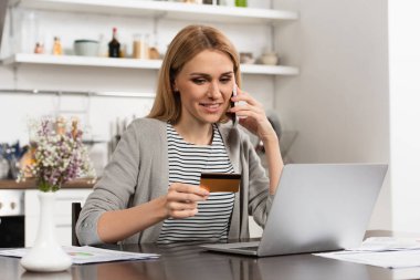 İnternetten alışveriş yaparken kredi kartı tutan ve cep telefonuyla konuşan mutlu bir kadın. 