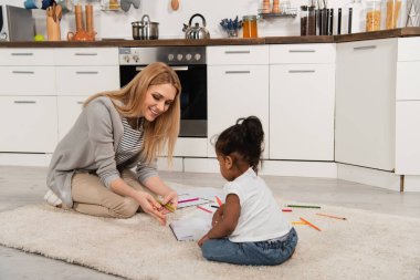 happy mother drawing with adopted african american girl while sitting on carpet  clipart