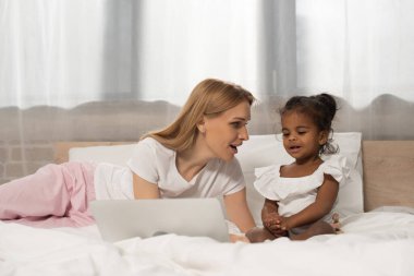 surprised mother and adopted african american girl watching movie on laptop in bedroom  clipart