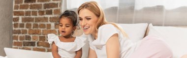happy mother and adopted african american girl watching movie on laptop in bedroom, banner clipart