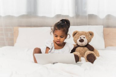 african american kid watching movie on laptop near soft toy  clipart