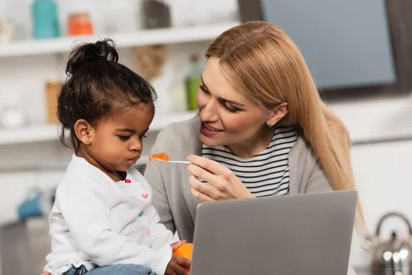 Šťastná Matka Krmení Adoptované Africké Americké Dcery Želé Blízkosti Notebooku — Stock fotografie