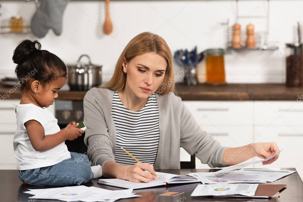 woman writing in notebook while working from home near adopted african american daughter