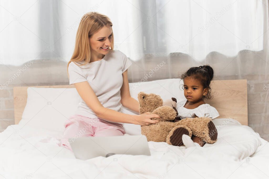 happy mother holding teddy bear near adopted african american kid watching movie on laptop in bedroom 