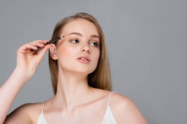 young woman applying brown eyebrow pencil and looking away isolated on grey clipart