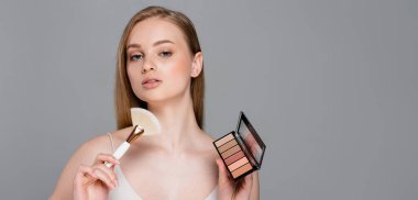young woman holding cosmetic brush and eye shadow and blush palette isolated on grey, banner clipart