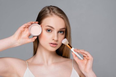 young woman holding cosmetic brush and face powder isolated on gray clipart