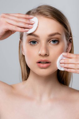 young woman with blue eyes removing makeup with cotton pads isolated on grey clipart