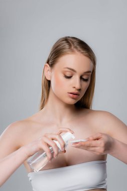 young woman with bare shoulders applying micellar water on cotton pad isolated on grey clipart