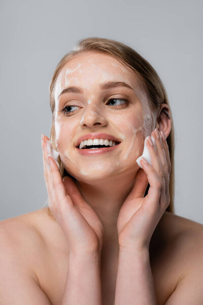 радостная молодая женщина с голубыми глазами и очищающей пеной на лице изолированы на сером