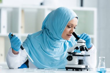 Türbanlı Arap bilim adamı mikroskoptan bakıyor ve elektronik pipeti tutuyor. 