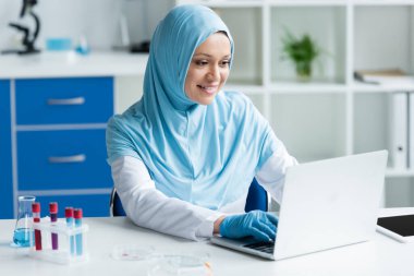 Test tüpleri, petri kapları ve dijital tabletin yanında dizüstü bilgisayar kullanan pozitif Arap bilim adamı 