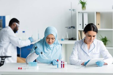 Test tüplerinin yanında elektronik pipet kullanan Arap bilim adamı ve laboratuarda dijital tabletle çalışan meslektaşı. 