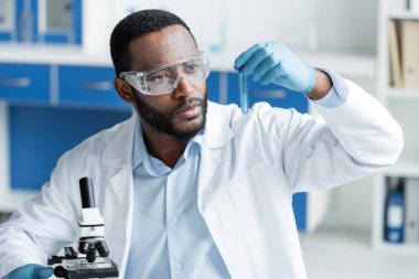 African american scientist in goggles holding test tube near microscope in laboratory  clipart