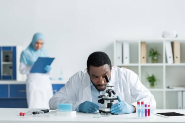 Beyaz önlüklü Afrikalı Amerikalı bilim adamı test tüpleri ve dijital tablet yakınındaki mikroskoptan bakıyor. 