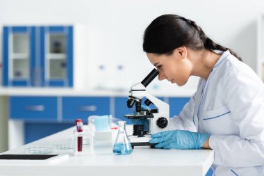 Side view of scientist in latex gloves using microscope near digital tablet with blank screen and equipment clipart
