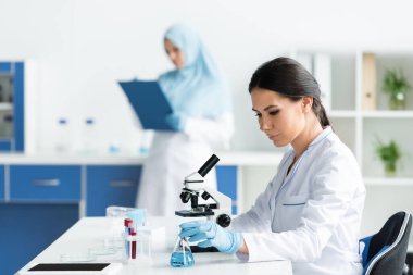 Tıbbi maskeli bilim adamı mikroskop ve test tüplerinin yanında matara alıyor. 