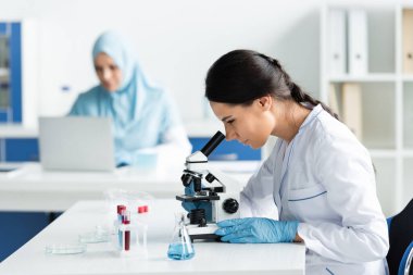 Lateks eldivenli bilim adamı, reaktör ve test tüpleriyle mikroskop yakınına bakıyor. 