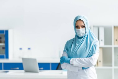 Tıbbi maskeli ve tesettürlü Müslüman doktor klinikte çapraz kollarla ayakta duruyor 