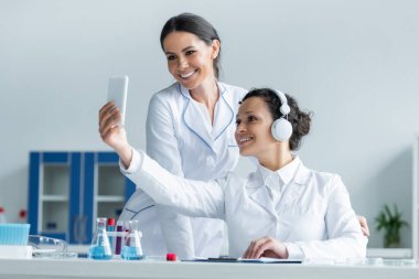 Kulaklıklı gülümseyen Afrikalı Amerikalı bilim adamı laboratuvarda iş arkadaşının yanında akıllı telefon kullanıyor. 